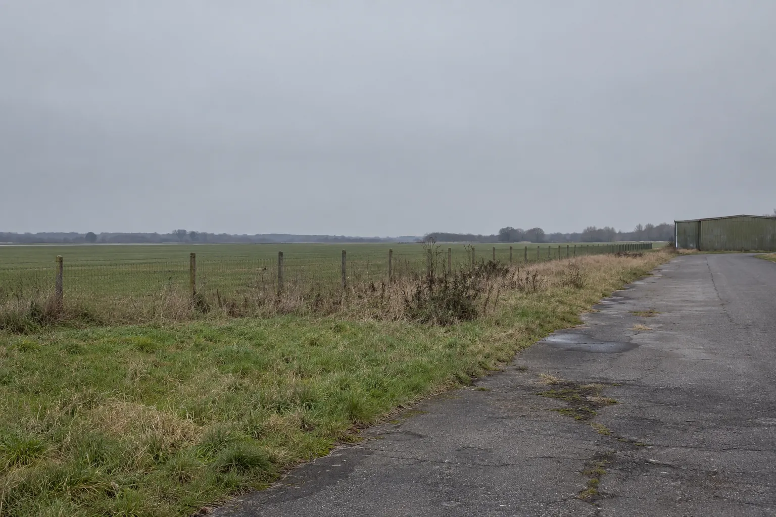 Een smalle asfaltweg langs een hek en grasland aan de rand van het vliegveld, met een loods in de verte.