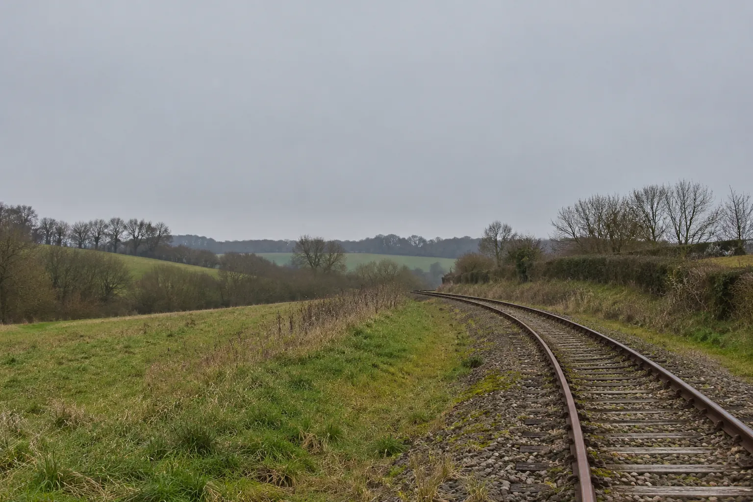 Een enkel spoor dat met een bocht door een groen heuveldal loopt, met heggen en kale bomen.