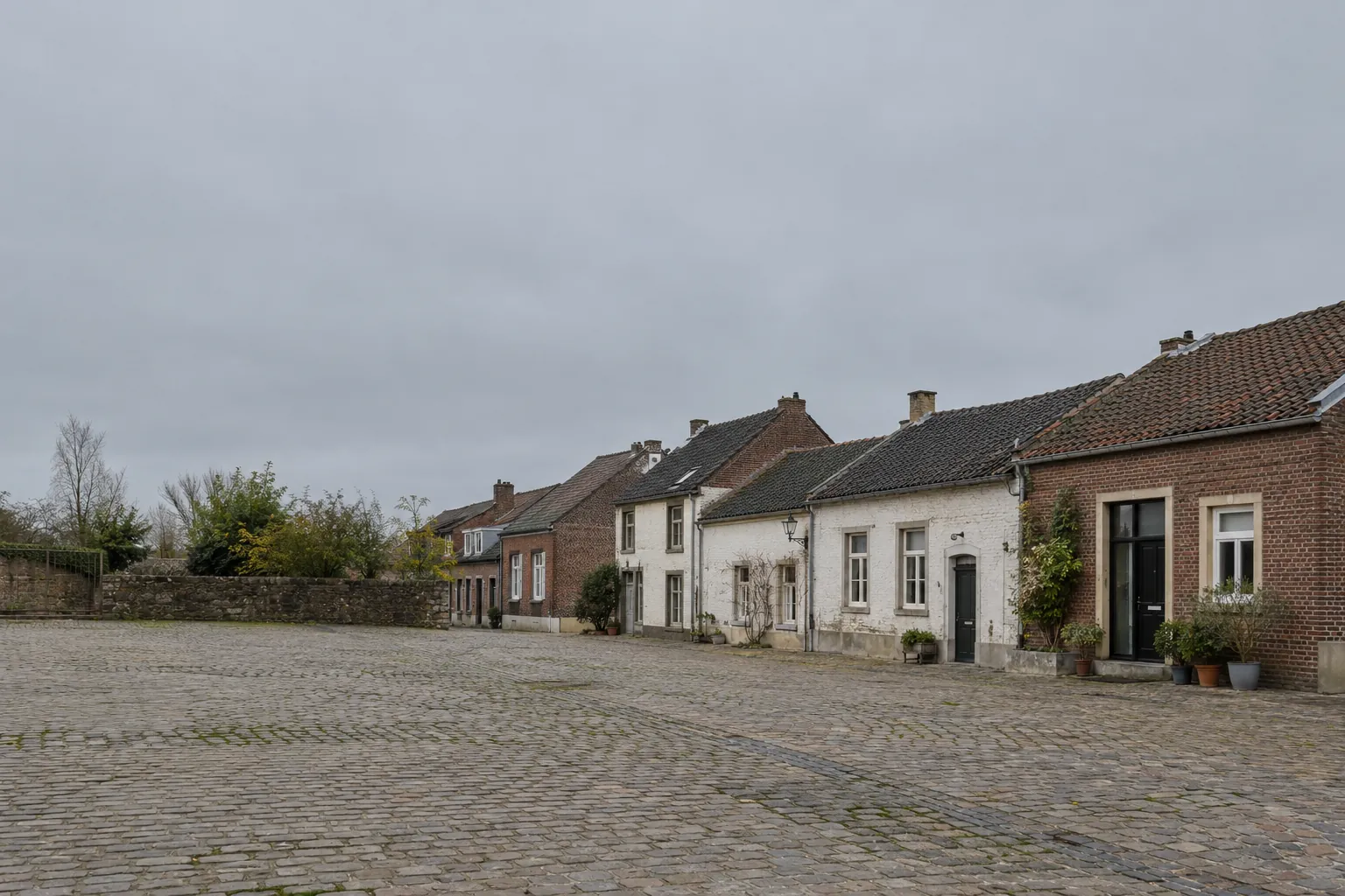 Een stille straat met kasseien en oude huizen van baksteen en wit pleisterwerk, met potplanten naast de voordeuren.