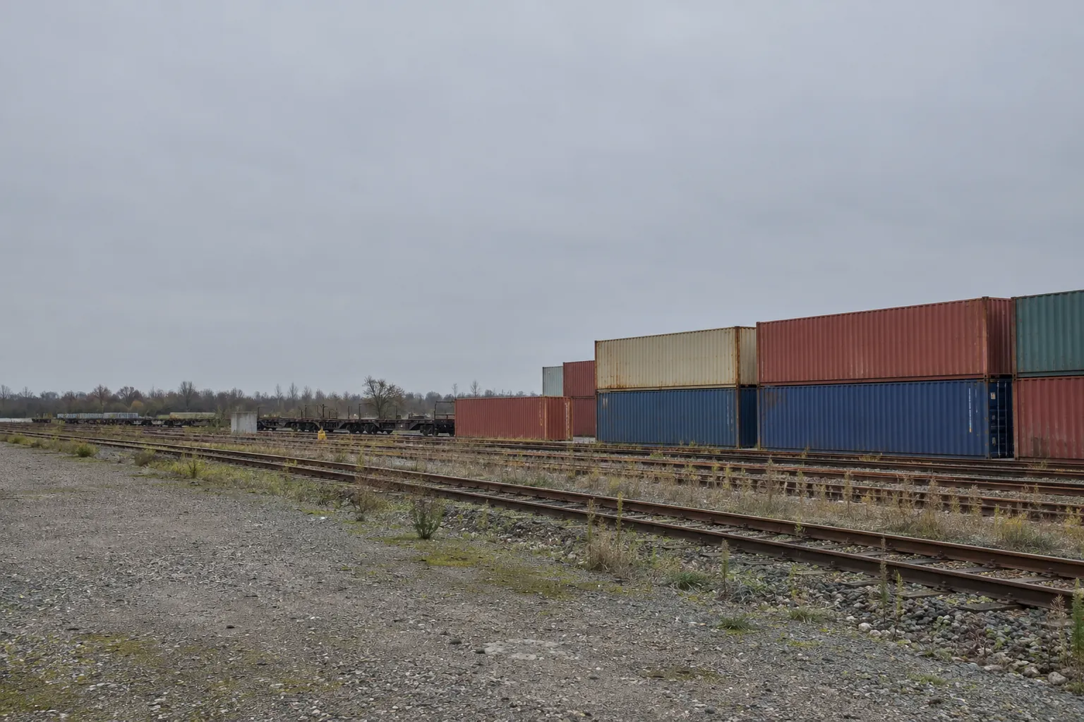 Een emplacement met sporen in onkruidig grind en gestapelde containers aan de rand van de stad.